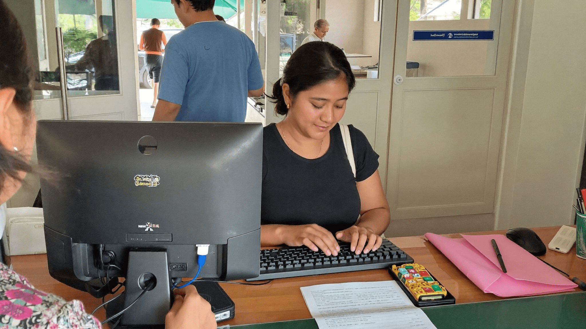 Reception in a modern Mumbai office, a compact desktop PC tucked behind the counter, receptionist working with a visitor