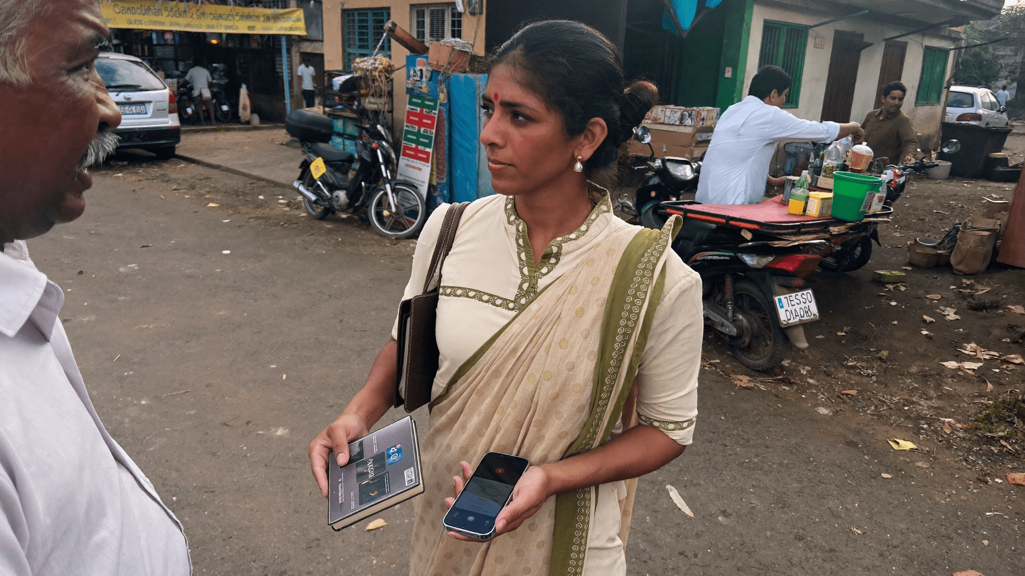 Pune field relationship manager in cream kurta on a neighbourhood street holding a corporate Android phone and a leather notebook in conversation with an MSME shopkeeper