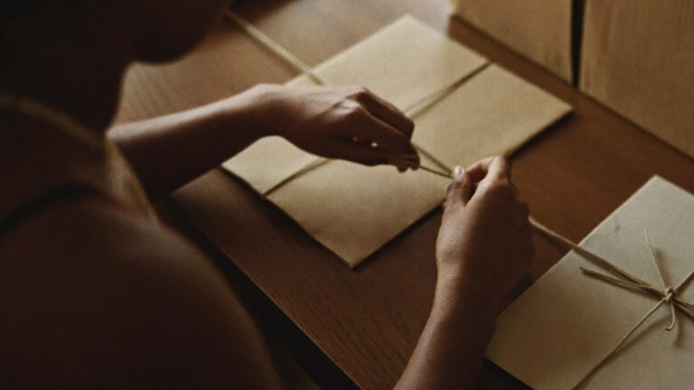 Hands of an Indian office coordinator tying jute twine around a plain kraft sleeve while packing low-budget corporate gifts on a wooden table