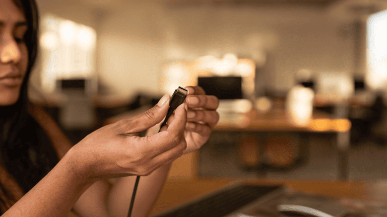 Hands of an Indian office IT lead holding a single cable connector before plugging it into a desk, with a blurred open-plan office behind