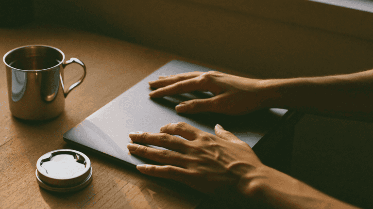 Hands of a young startup founder resting on a closed laptop beside a steel tumbler of chai in a small Indian software office