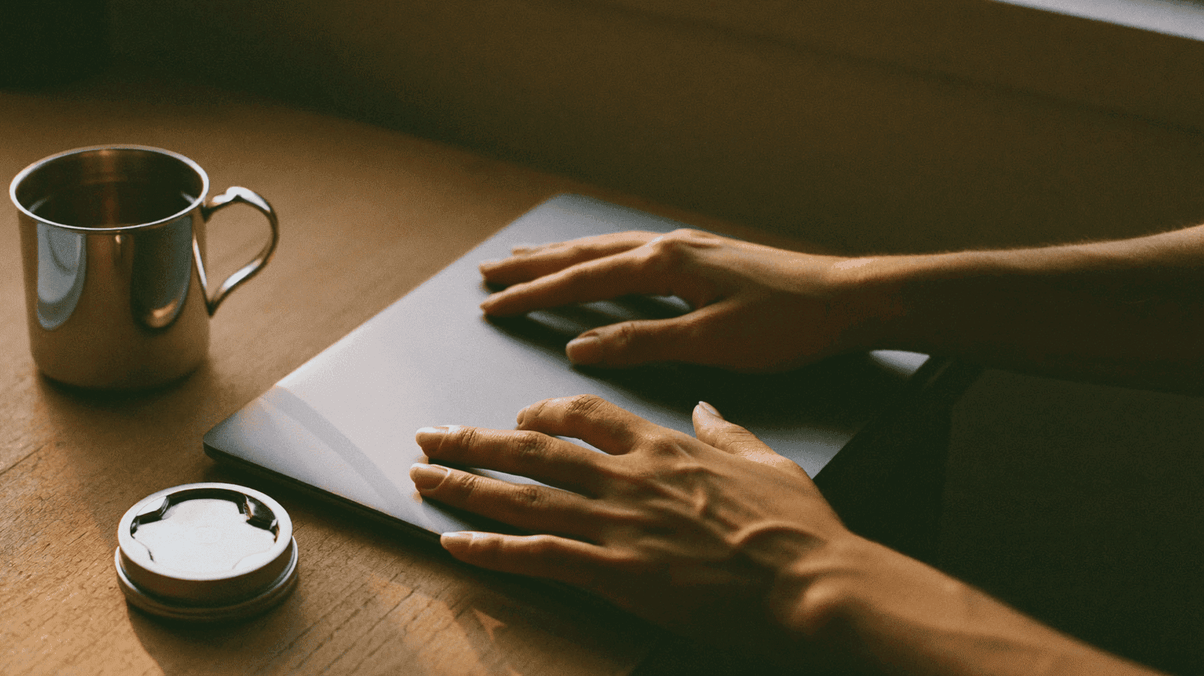 Hands of a young startup founder resting on a closed laptop beside a steel tumbler of chai in a small Indian software office