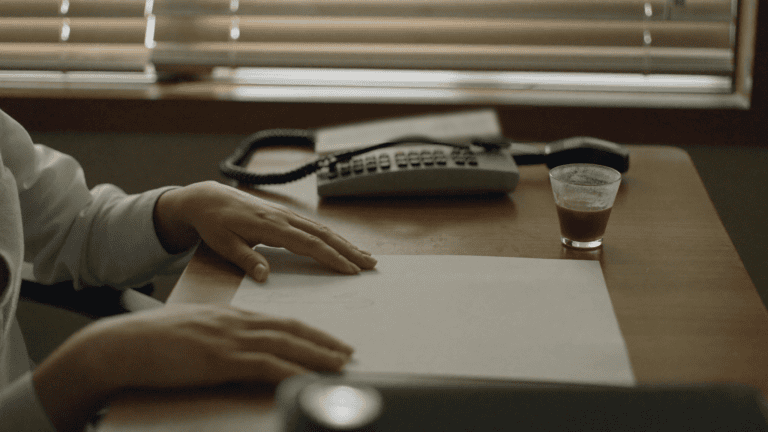 Hands of a compliance lead resting on a blank sheet of paper beside a landline phone and a glass of chai in a Jaipur office