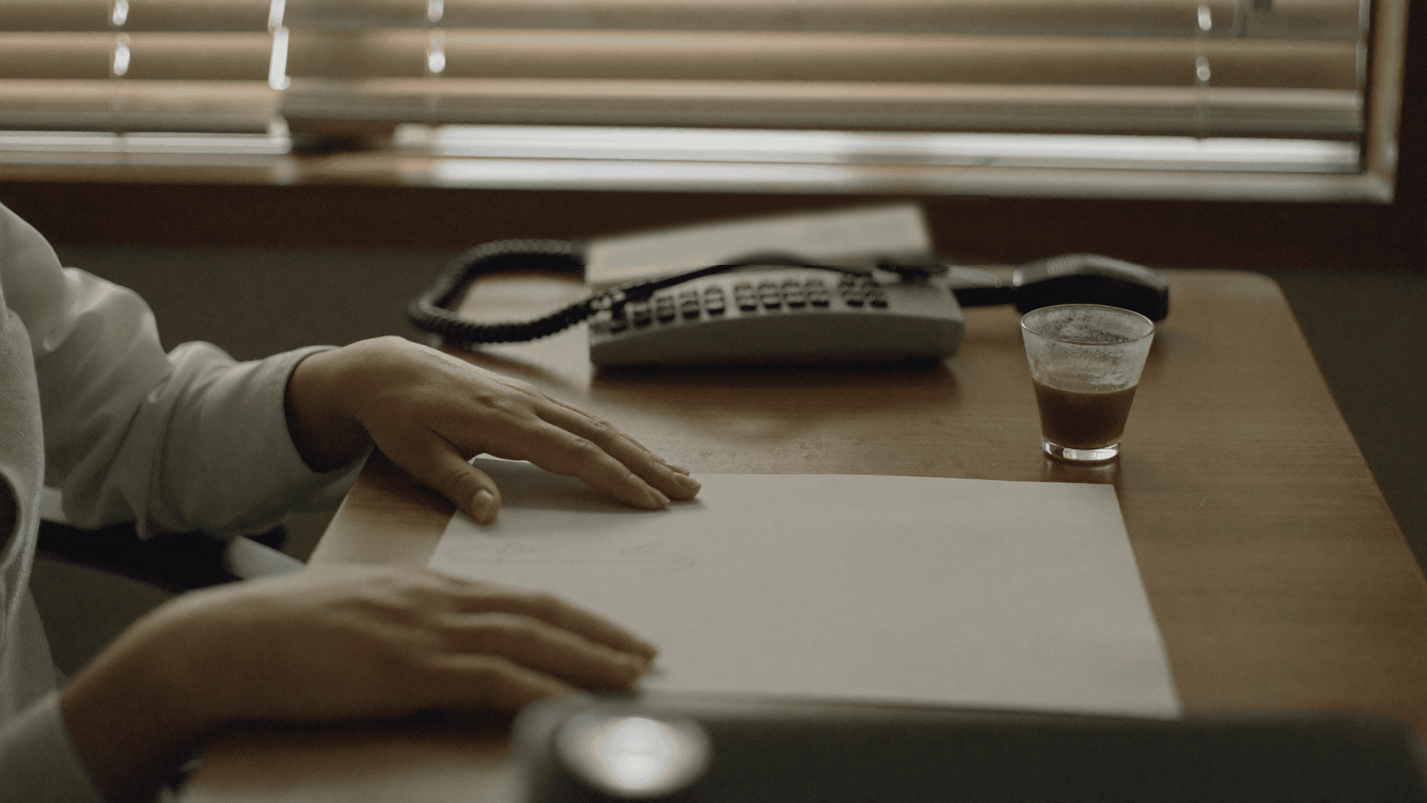 Hands of a compliance lead resting on a blank sheet of paper beside a landline phone and a glass of chai in a Jaipur office