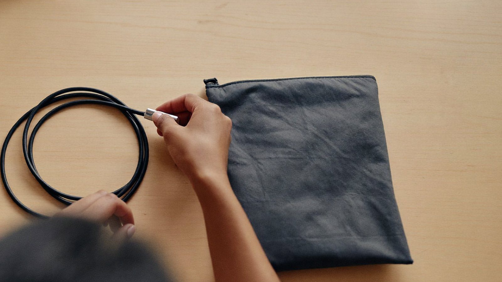 Hands of an Indian HR coordinator coiling a charging cable next to a plain fabric pouch while assembling an employee welcome kit on a desk