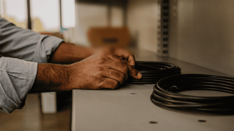 Hands of an Indian IT administrator lifting a coiled power cable off a bare metal shelf during a physical device count in a company store room