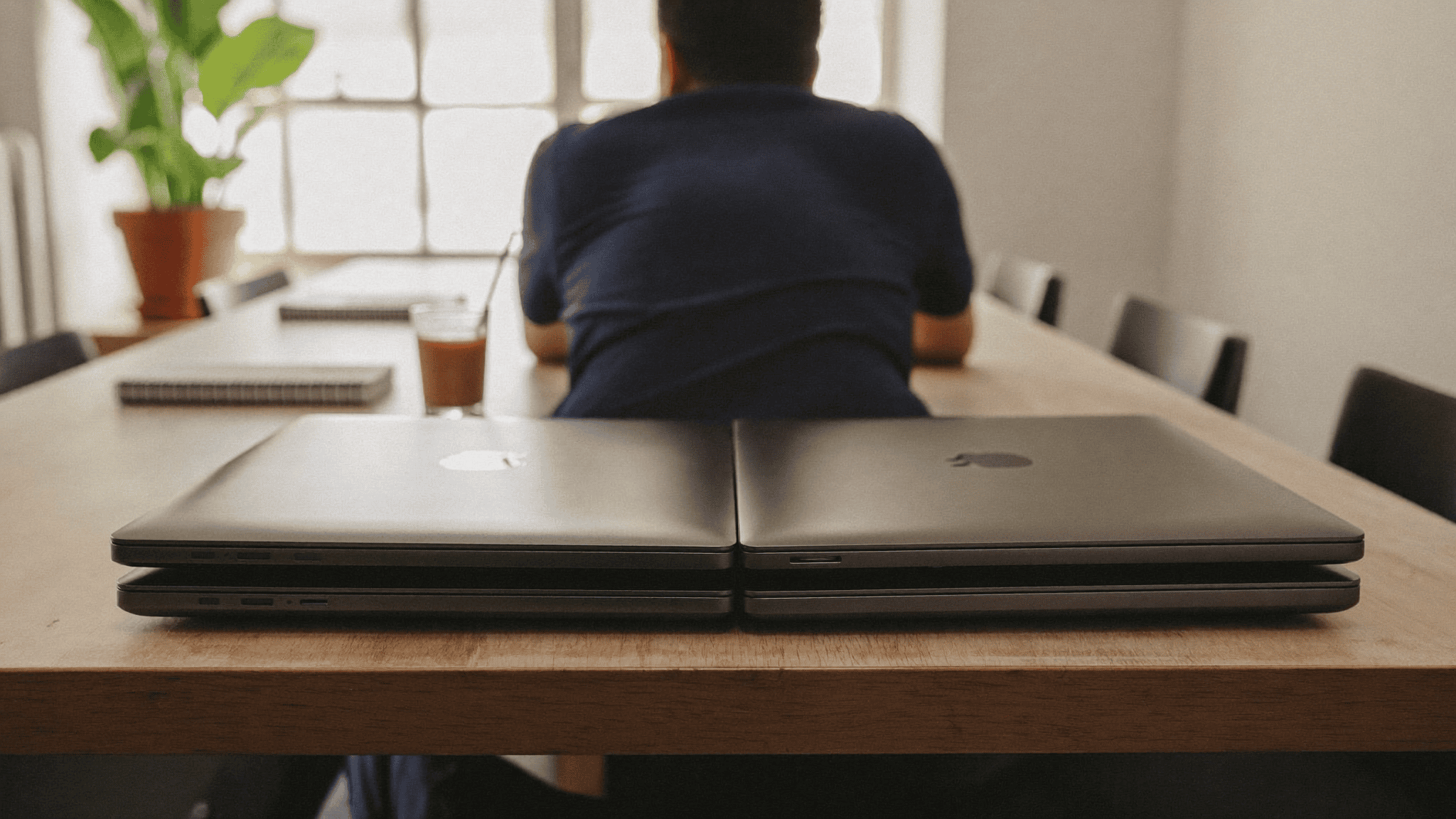 An IT manager sitting at a boardroom table in an Indian office with two stacks of closed company laptops in front of him during an Apple device management review