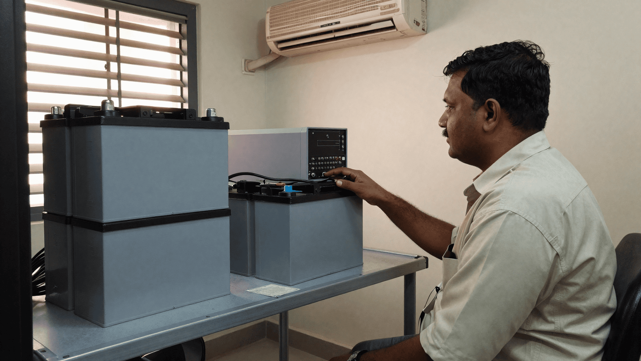 A facilities manager in a small first-floor Coimbatore office server cabin inspects a rack UPS beside two clearly different battery packs: a larger stack of traditional lead-acid style blocks on the left and a compact modern lithium module on the right. Sirius Star.