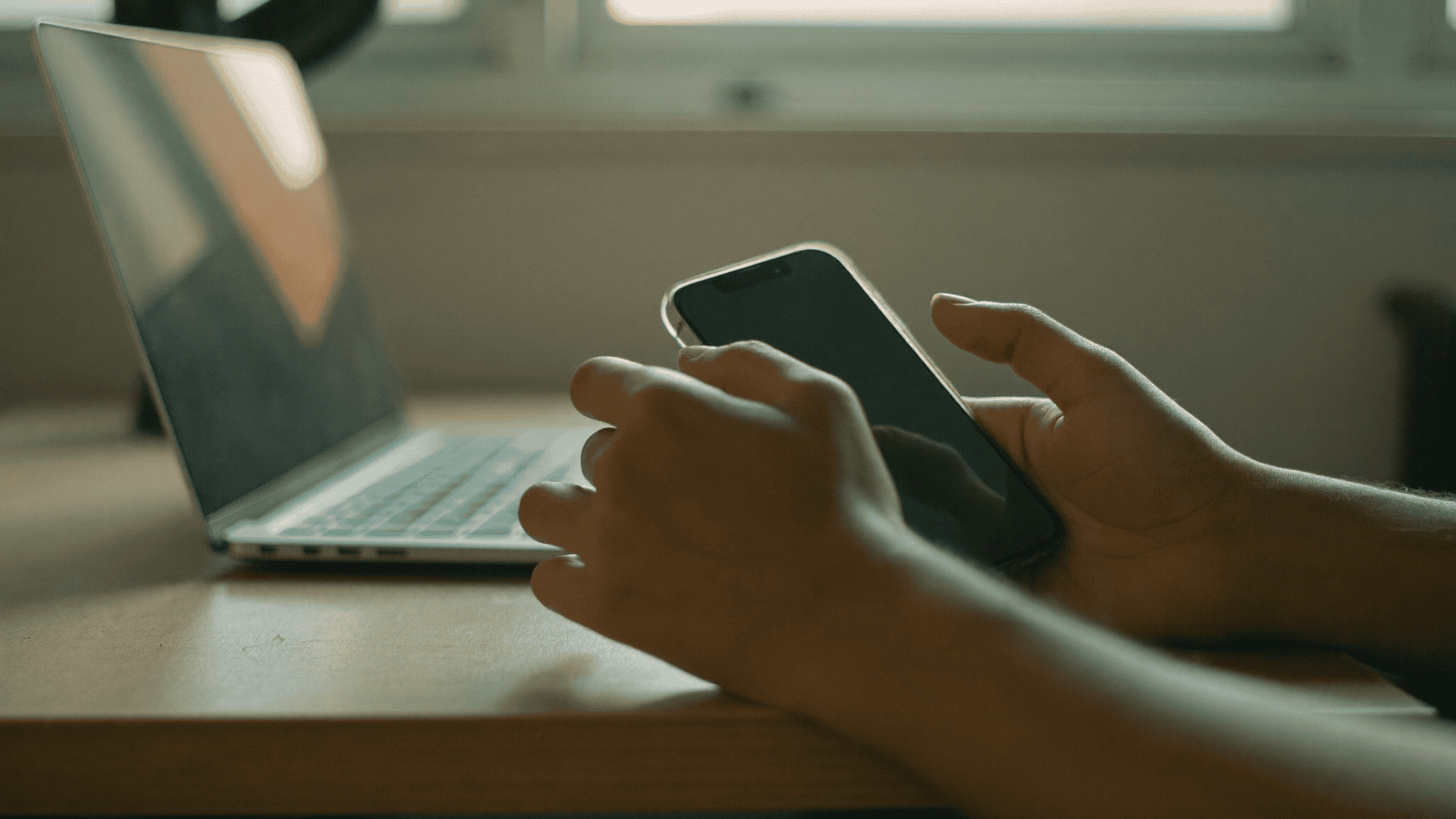Hands holding a phone with a dark screen beside an open laptop on a wooden desk in an Indian office, late afternoon light