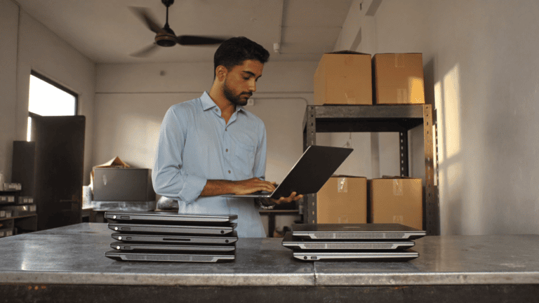 An IT administrator in an Indian company store room checking a business laptop, with two separate stacks of closed office laptops on the bench in front of him during a four-year fleet review