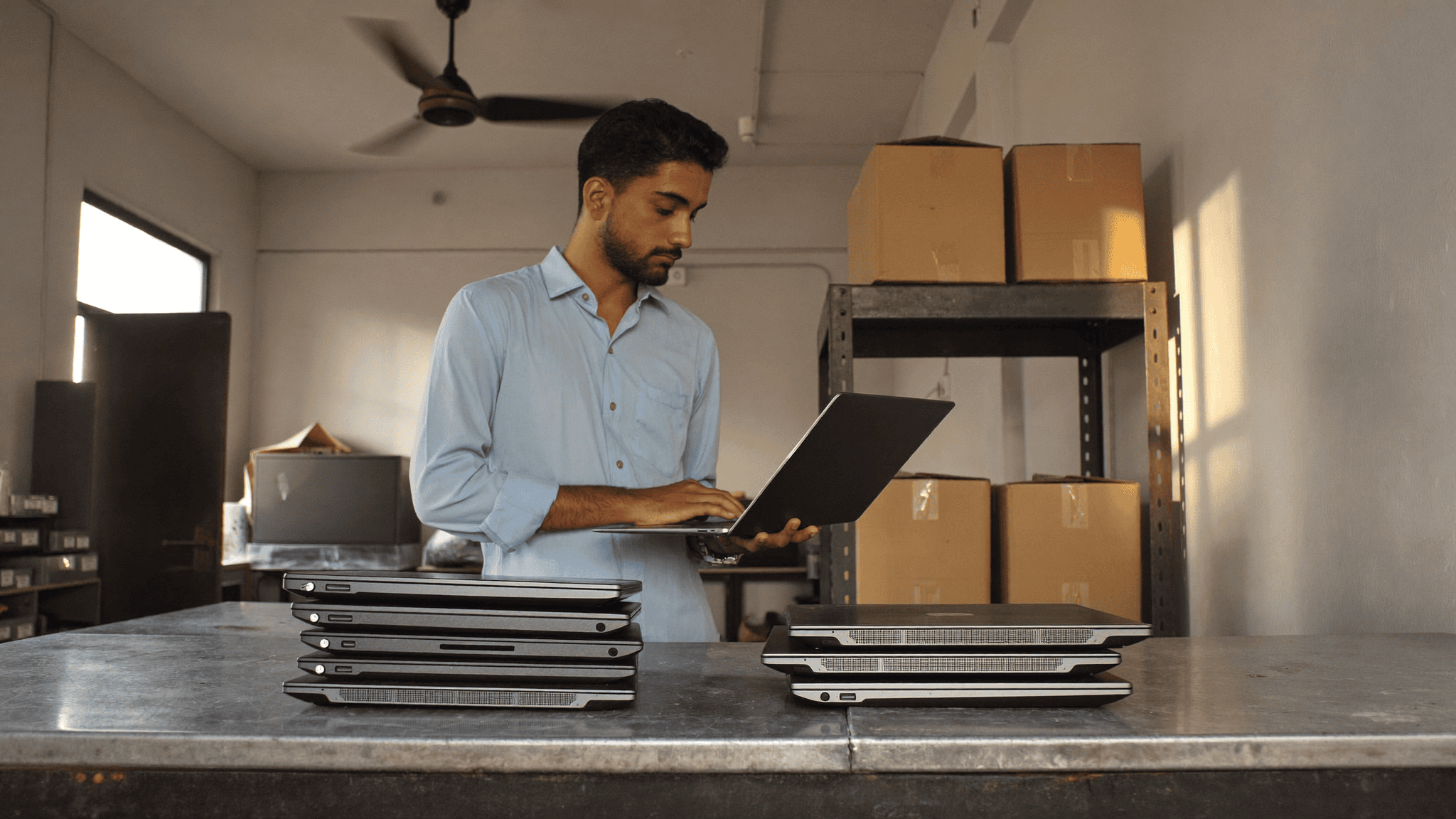 An IT administrator in an Indian company store room checking a business laptop, with two separate stacks of closed office laptops on the bench in front of him during a four-year fleet review