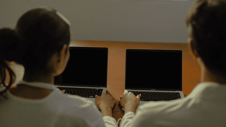 An IT security consultant and a business manager seen from behind, comparing two laptops with blank screens at a desk in a mid-size Navi Mumbai office